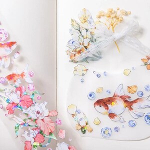 K&ouml;nnte beinhalten: Ein Blatt mit transparenten Aufklebern mit verschiedenen bunten Illustrationen. Die Aufkleber zeigen Goldfische, Blumen und Blasen. Die Aufkleber sind perfekt, um Tageb&uuml;chern, Planern und anderen Bastelprojekten einen Hauch von Verspieltheit zu verleihen.