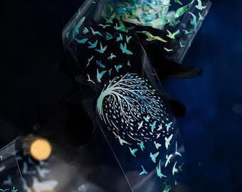 May include: Clear, transparent tape with a design of a tree and flying birds in shades of blue and green. The tape is curled, showing the design from different angles against a dark background.