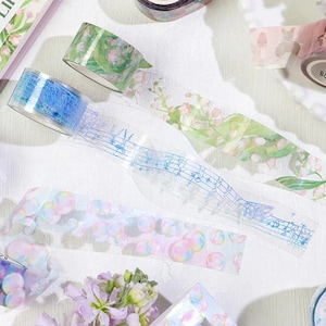 May include: Assortment of decorative washi tapes with various designs.  The tapes feature a floral pattern, musical notes, and a bubble pattern.  The tapes are transparent and have a subtle shimmer.