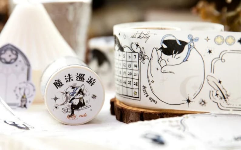 May include: Two rolls of decorative washi tape with black and white illustrations. One roll features a cat sleeping on a crescent moon with stars and the text "one cat kitty". The other roll features a cat wearing a witch's hat with the text "魔法 巡游" and "really cute".