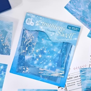 May include: A collection of blue and white decorative paper sheets with a celestial theme. The package reads "Miss Time Luminous Shower Swell" with a design of stars and water. Other sheets feature water and wave patterns.