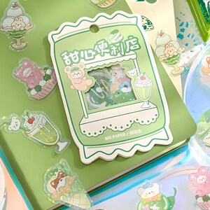 May include: A collection of colorful, transparent stickers featuring cute cartoon bears, desserts, and a green shop design. The stickers include images of sundaes, ice cream cones, and a shop with the text "Sweet Convenience Store".