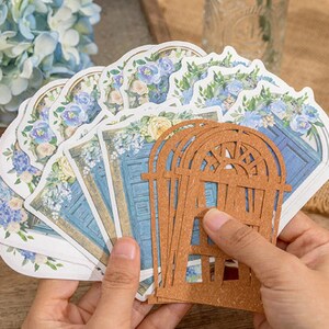 May include: A set of decorative paper die cuts featuring blue doors with floral accents. The die cuts are arranged in a fan shape and are held in a hand. A brown wooden die cut of a window is in front of the paper die cuts.