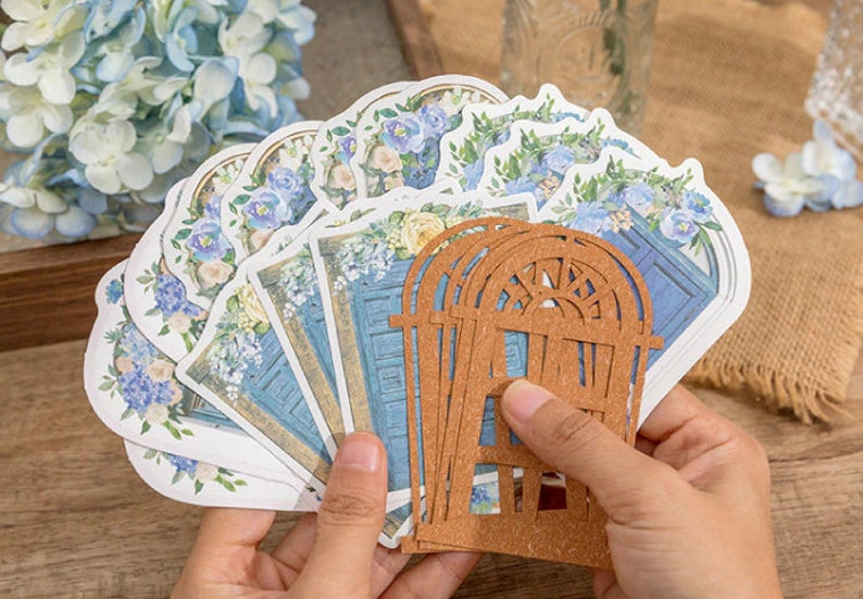 May include: A set of decorative paper die cuts featuring blue doors with floral accents. The die cuts are arranged in a fan shape and are held in a hand. A brown wooden die cut of a window is in front of the paper die cuts.