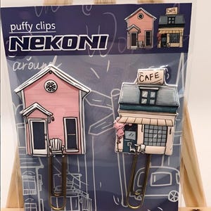 May include: Two puffy paper clips shaped like buildings. One is a pink house with a white door and a rocking chair on the porch. The other is a blue and white cafe with a sign that says "Cafe". The clips are on a blue and white background with the text "puffy clips NEKONI around".