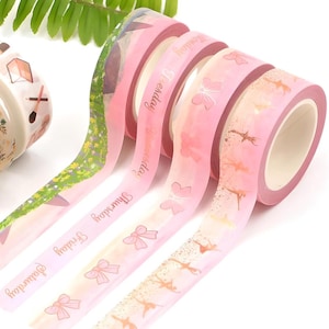 May include: Four rolls of pink washi tape with gold foil accents. The tape features designs of bows, butterflies, and dancers. The text on the tape reads "Dreamland", "Thursday", "Happy Birthday", and "Be Happy".