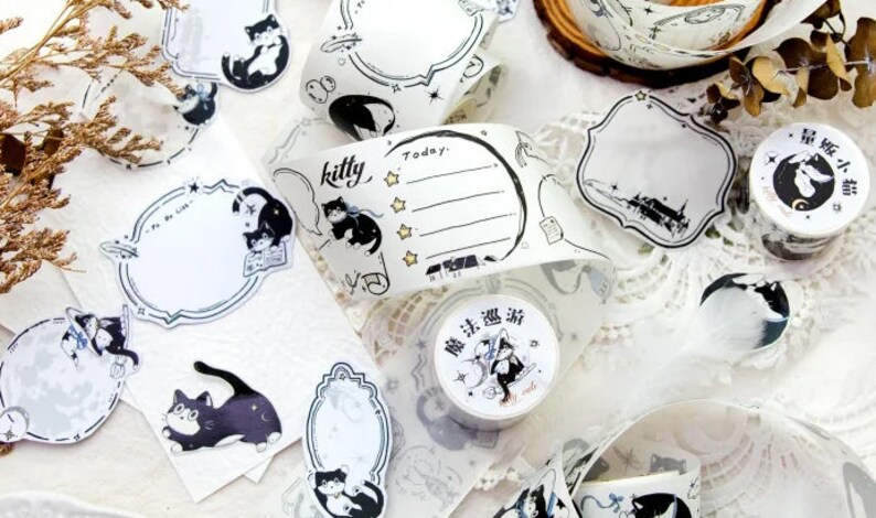 May include: Assortment of decorative washi tapes featuring black and white illustrations of cats, stars, and moons. The tapes are on a white background with a lace overlay. The text "kitty" and "Today" are visible on one of the tapes.