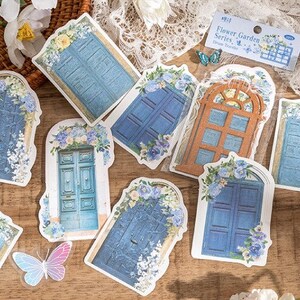 May include: A set of nine decorative stickers featuring blue and brown doors with floral accents. The stickers are part of a "Flower Garden Series" and are designed for use in a traveler's journal.