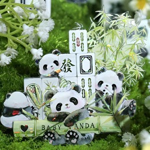 May include: A charming display of panda bear figurines with black and white markings, set amongst bamboo and Mahjong tiles. The words "BABY PANDA" are displayed on a bamboo stalk. The background features greenery and white flowers.