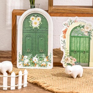 May include: Two green arched doors with floral details and white flowers. The doors are on a wooden shelf with a white picket fence and two white ceramic sheep figurines in front of them.