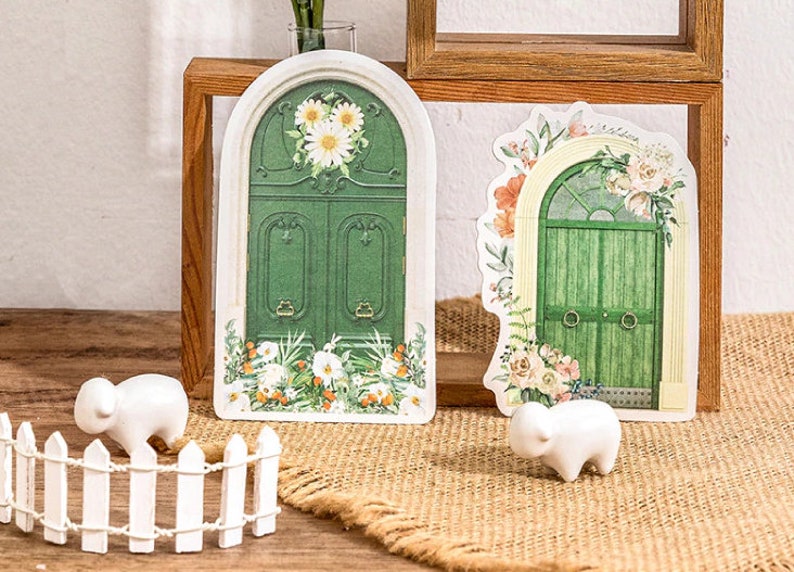 May include: Two green arched doors with floral details and white flowers. The doors are on a wooden shelf with a white picket fence and two white ceramic sheep figurines in front of them.