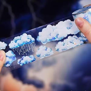 May include: A roll of clear washi tape with a repeating pattern of blue and white clouds. Some of the clouds have raindrops falling from them.