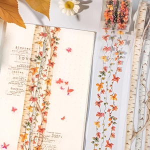 May include: Washi tape with a design of autumn leaves in orange, red, and yellow hues. The tape has a transparent background, giving the impression of floating leaves. A notebook with text and birch tree branches are also visible in the image.