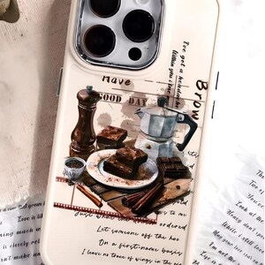 May include: A white phone case with a watercolor design of a coffee pot, chocolate squares, and a plate of brownies. The text "Have a Good Day" is written on the case.