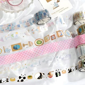 May include: Assortment of colourful washi tapes with various designs, including geometric patterns, animals, and illustrations. The tapes are arranged on a white surface with the text "Energy and persistence conquer all things".