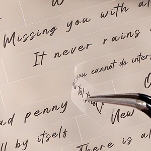 May include: A close-up of a clear sticker with the text "It never rains but it pours" written in black ink. The sticker is being peeled off of a white surface with the text "Missing you with all" and "ad penny" visible.
