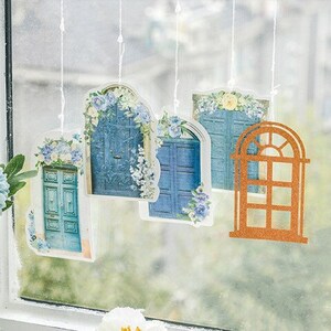 May include: Five decorative door-shaped ornaments hanging from a window. The ornaments are made of paper and feature blue doors with floral details. One ornament is a brown window frame.