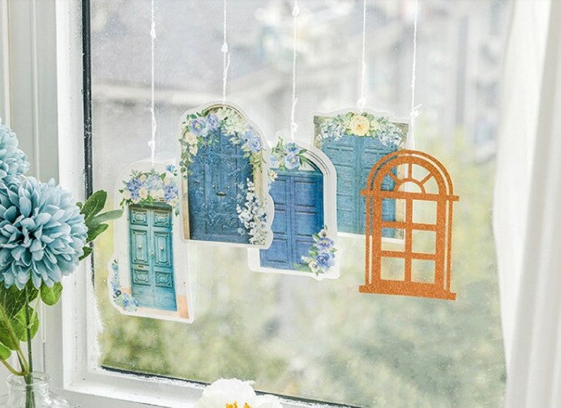May include: Five decorative door-shaped ornaments hanging from a window. The ornaments are made of paper and feature blue doors with floral details. One ornament is a brown window frame.