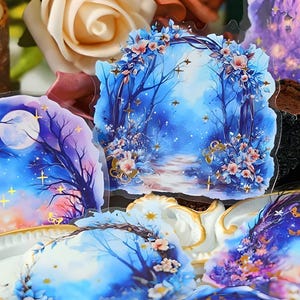 May include: A selection of fantasy-themed stickers with illustrations of moonlit forests, starry skies, and floral arches. The stickers have a watercolour effect with shades of blue, purple, and pink, and are displayed on a decorative tray.
