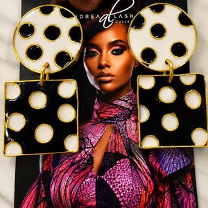 May include: A pair of large statement earrings featuring a gold-toned metal frame. Each earring consists of a circular top piece with a white background and black polka dots, connected to a square bottom piece with a black background and white polka dots.