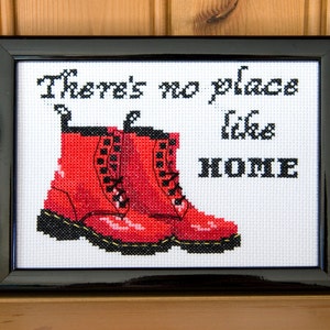 Puede incluir: Bordado de punto de cruz en un marco negro con botas rojas Doc Martens y el texto "There's no place like HOME".