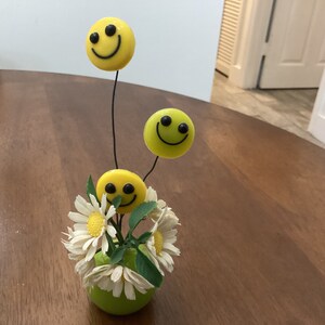 May include: A green ceramic vase with white daisies and three yellow smiley faces on wire stems.