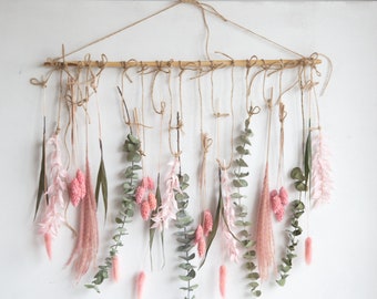 May include: A wall hanging decoration made from a wooden rod, twine, and dried flowers. The arrangement includes pink, green, and beige dried flowers and foliage. Purple flowers hang from above. A natural, bohemian style home decor piece.