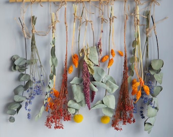 May include: A wall hanging made of dried flowers and greenery. The flowers are arranged in a row on a wooden dowel and are tied with twine. The flowers are a variety of colors, including orange, yellow, and purple. The greenery is a variety of shades of green.