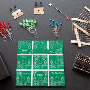 May include: A green circuit board with the text "Crafty Electron" and "Tic-Tac-Toe" printed on it. The board has six sections, each with a different design. There are also various electronic components, including resistors, LEDs, and transistors, scattered around the board.