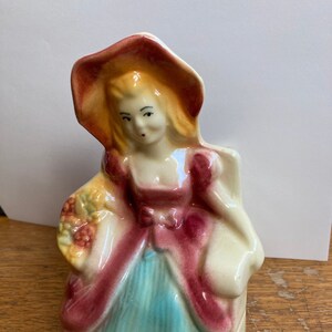 May include: A ceramic figurine of a woman wearing a red and yellow hat, a red dress with a blue skirt, and holding a bouquet of flowers. The figurine is a planter.