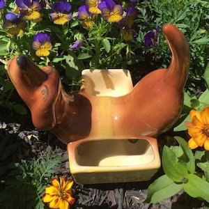 May include: A brown and tan ceramic dachshund planter with a small, rectangular planter inside. The planter is sitting on a bed of brown mulch and surrounded by purple and yellow flowers.