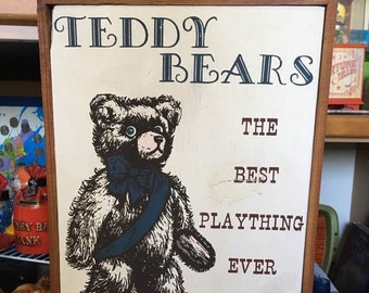 May include: A vintage sign with a brown wooden frame and white background. The sign reads "Teddy Bears The Best Plaything Ever Invented". A cartoon teddy bear with blue eyes and a blue sash is pictured on the sign.