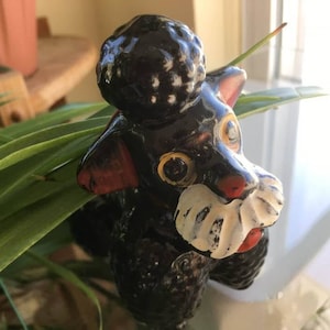 May include: Black and white ceramic poodle figurine with yellow eyes and a white mustache. The figurine is sitting on a green plant.