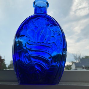 May include: A cobalt blue glass bottle with an oval shape and a narrow neck. The bottle features a raised floral design on the front and back. The bottle is sitting on a white windowsill.