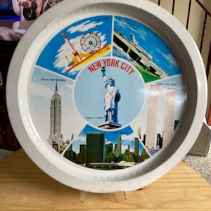 May include: A round white metal serving tray featuring a central image of the Statue of Liberty with the text New York City. Surrounding the center are four panels depicting the Empire State Building, the World Trade Center, a carnival, and a bridge.