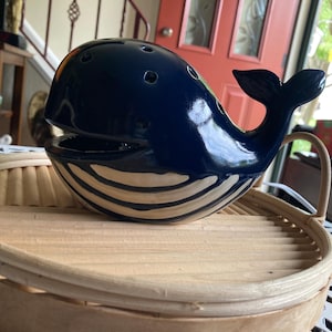 May include: A glossy dark blue ceramic whale figurine with a wide open mouth and a series of small circular holes along the top of its head. The whale features a cream-colored underbelly with horizontal ridges and a raised tail fin.