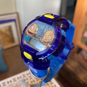 May include: A blue digital wristwatch featuring characters from the animated series Rugrats. The watch has a blue band and a clear face with a cartoon image. Yellow accents are on the watch face.