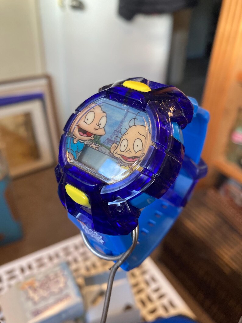 May include: A blue digital wristwatch featuring characters from the animated series Rugrats. The watch has a blue band and a clear face with a cartoon image. Yellow accents are on the watch face.