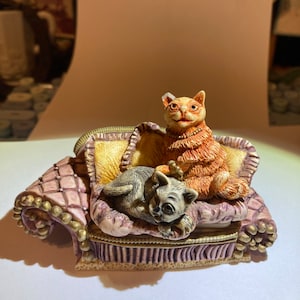 May include: A ceramic figurine of two cats sitting on a purple and gold couch. The cat on the left is gray and white, while the cat on the right is orange and white.