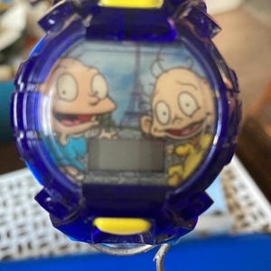 May include: A blue and yellow digital watch featuring cartoon characters. The watch face displays two characters with the Eiffel Tower in the background. The watch has a blue bezel and a digital display.