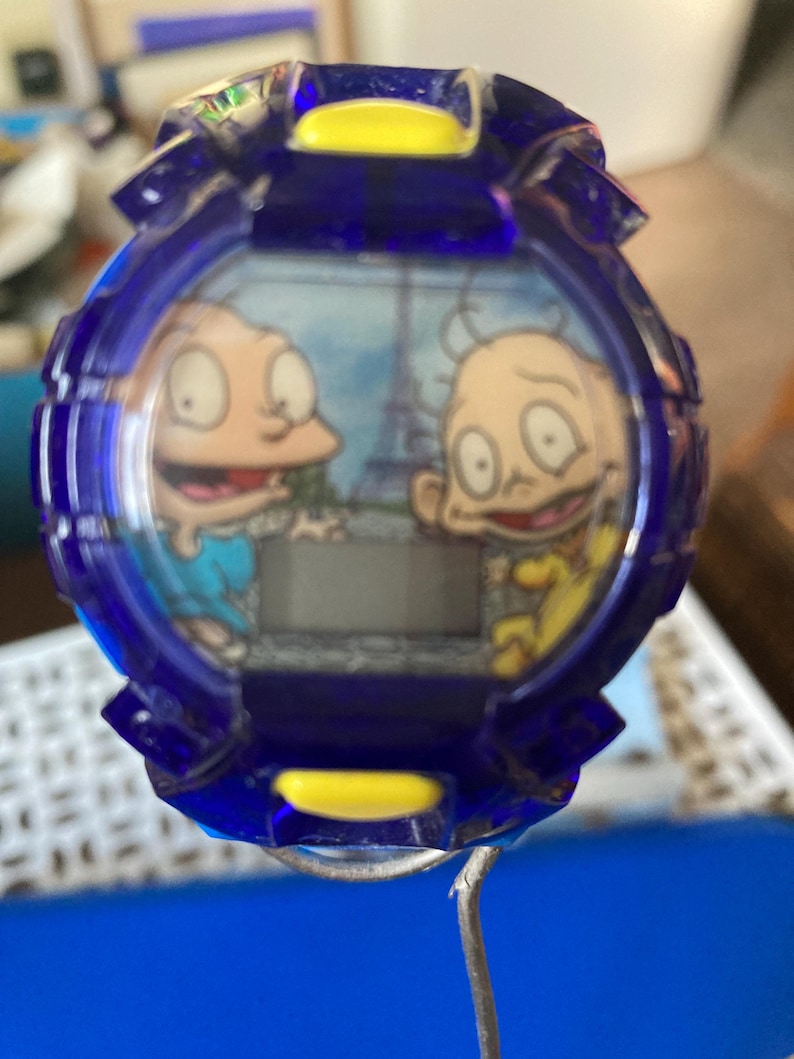 May include: A blue and yellow digital watch featuring cartoon characters. The watch face displays two characters with the Eiffel Tower in the background. The watch has a blue bezel and a digital display.