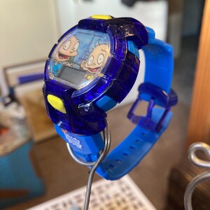May include: A blue and yellow digital wristwatch featuring cartoon characters. The watch has a blue band and a dark blue face with a digital display. The watch is held up by a metal wire.