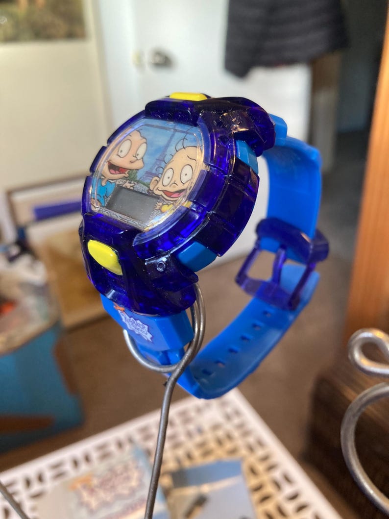 May include: A blue and yellow digital wristwatch featuring cartoon characters. The watch has a blue band and a dark blue face with a digital display. The watch is held up by a metal wire.