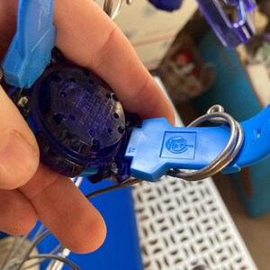 May include: A close-up of a blue and purple toy watch. The watch has a blue strap and a dark purple face. The watch is held together with a silver metal ring. The watch has a logo on the blue clasp.