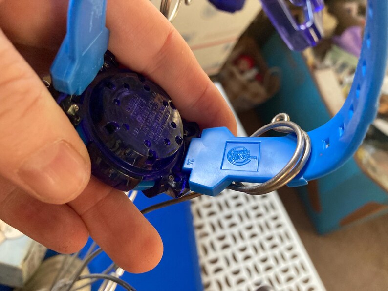 May include: A close-up of a blue and purple toy watch. The watch has a blue strap and a dark purple face. The watch is held together with a silver metal ring. The watch has a logo on the blue clasp.