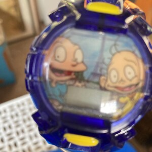 May include: A blue and yellow watch featuring a cartoon image of two characters. The watch has a round face with a blue bezel and a yellow accent. The characters are depicted in a cartoon style, with large eyes and simple features. The watch appears to be a vintage or collectible item.