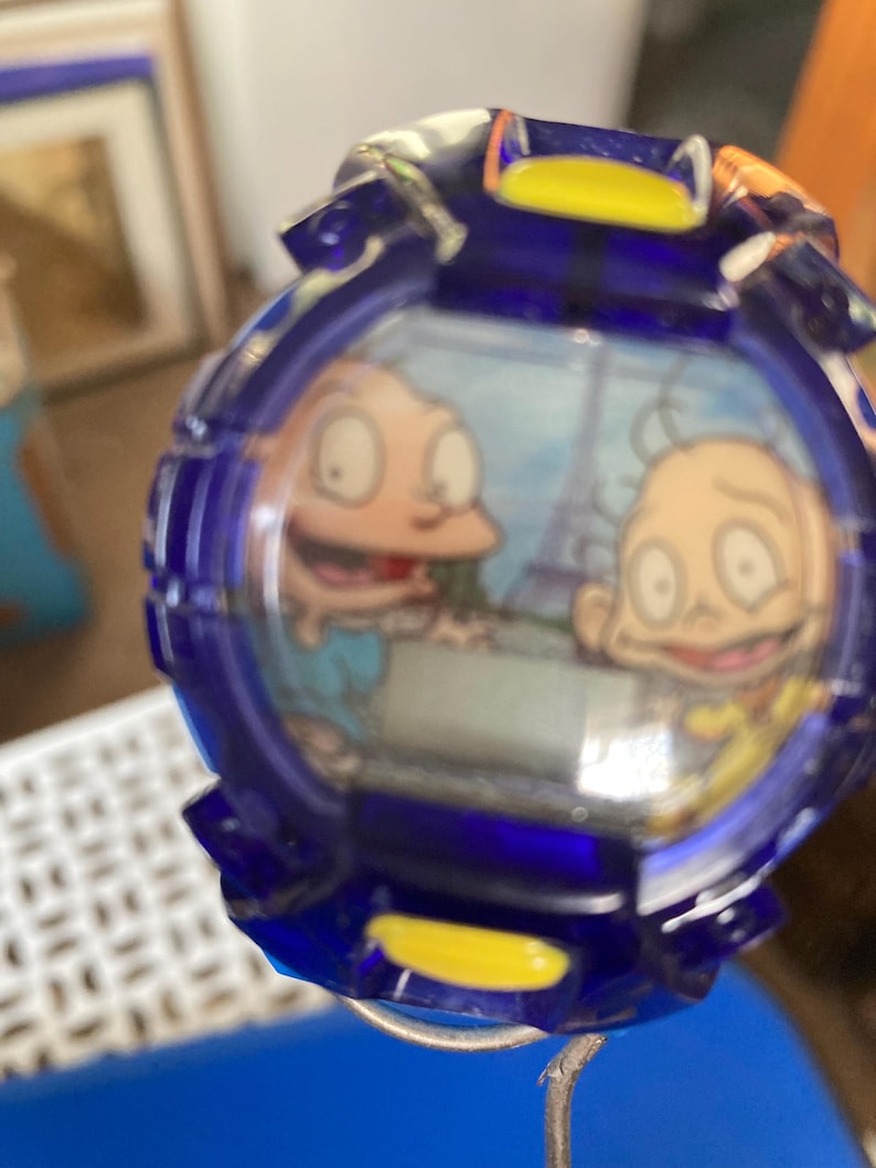 May include: A blue and yellow watch featuring a cartoon image of two characters. The watch has a round face with a blue bezel and a yellow accent. The characters are depicted in a cartoon style, with large eyes and simple features. The watch appears to be a vintage or collectible item.