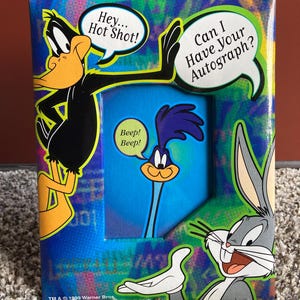 May include: A rectangular picture frame featuring Daffy Duck, Bugs Bunny, and the Road Runner. Daffy Duck says, Hey... Hot Shot!, while the Road Runner says, Beep! Beep!, and Bugs Bunny asks, Can I Have Your Autograph? The frame has a colorful background.
