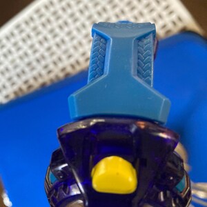 May include: A close-up of a blue and purple toy. The top portion is a solid blue with a textured design. The lower portion is a translucent purple with a yellow button and silver accents. The toy is set against a blue background.