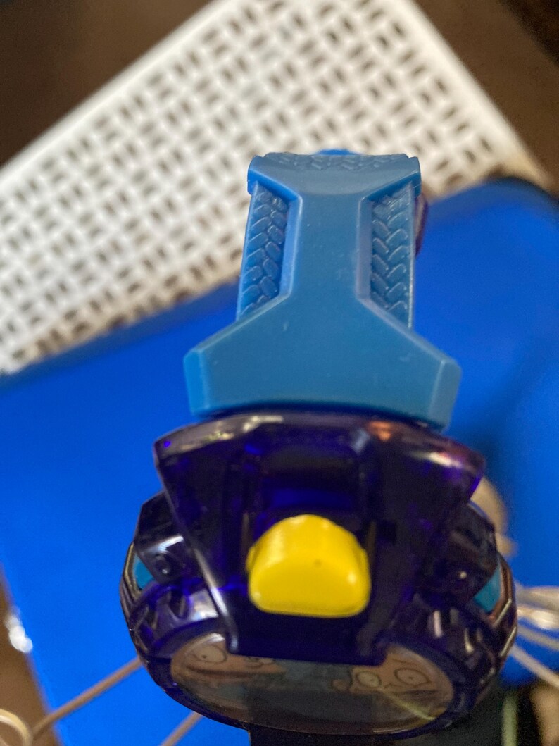 May include: A close-up of a blue and purple toy. The top portion is a solid blue with a textured design. The lower portion is a translucent purple with a yellow button and silver accents. The toy is set against a blue background.